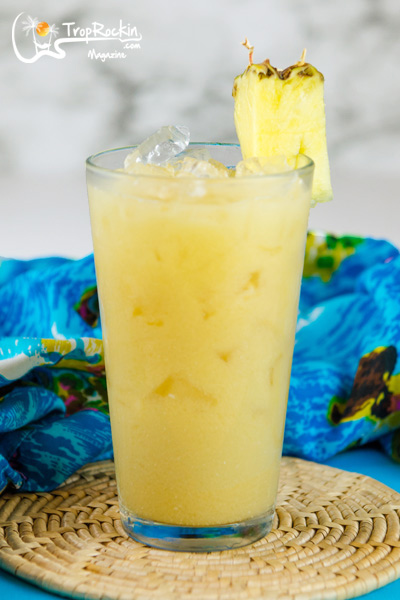 Painkiller Drink with Pineapple Wedge Garnish sitting on kitchen couner with tropical blue fabric background