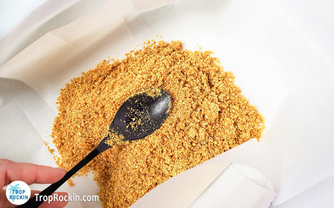 Pressing graham cracker crumbs with a spoon in a prepared baking pan lined with parchment paper.
