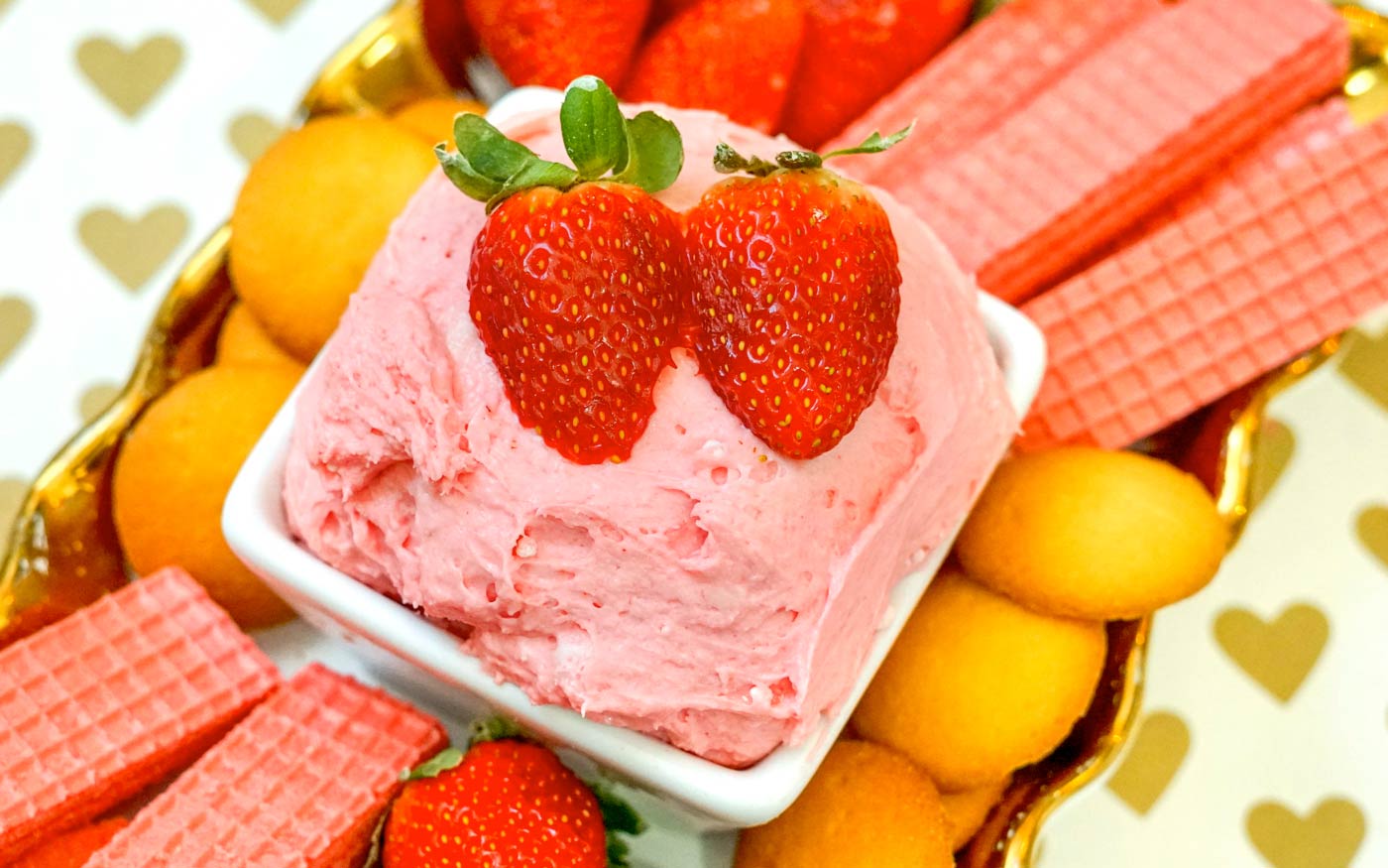 Pink Strawberry Fruit Dip in a white bowl with two Strawberries on top with a platter of strawberry wafers, nilla wafers and fresh strawberries.
