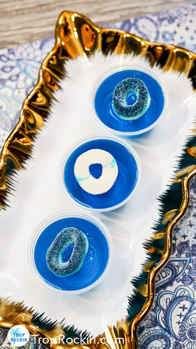 Three blue raspberry jello shots with blue and white gummies as garnish displayed on a gold and white serving dish.
