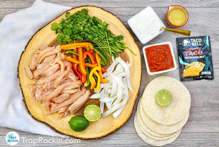 Recipe ingredients including sliced chicken, peppers, onions, limes, taco seasoning and traditional toppings of sour cream, salsa and cilantro on a wood table.