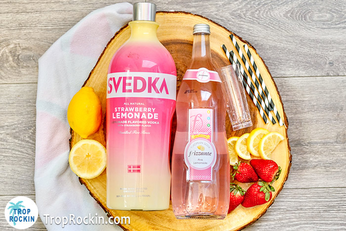 Strawberry lemonade vodka shots ingredients: Svedka Strawberry Lemonade Vodka bottle and pink lemonade italian soda bottle with sliced lemons and fresh strawberries.