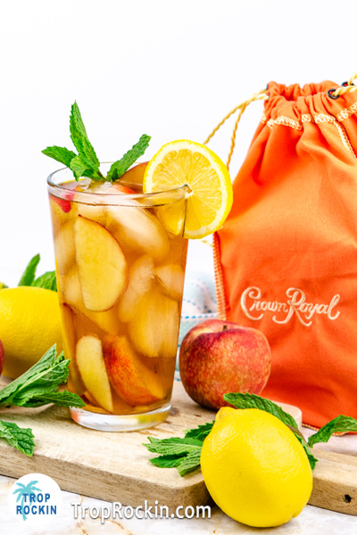 Crown Peach Tea drink on wood cutting board with peaches and lemons with Crown Royal Peach whisky bottle in the background.