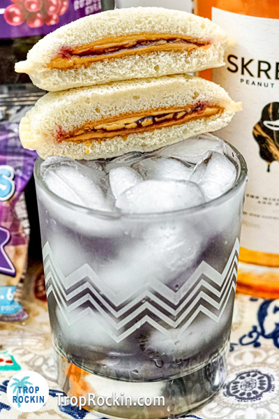 Upclose photo of this Peanut Butter Whiskey Drink with mini peanut butter and jelly sandwiches for garnish.Skrewball Peanut Butter Whiskey bottle in background.