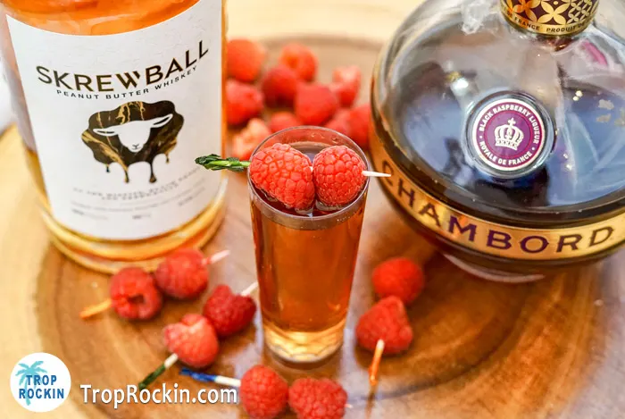 Close up view of Skrewball Whiskey and Chambord bottles with a peanut butter and jelly shot topped with fresh raspberries.
