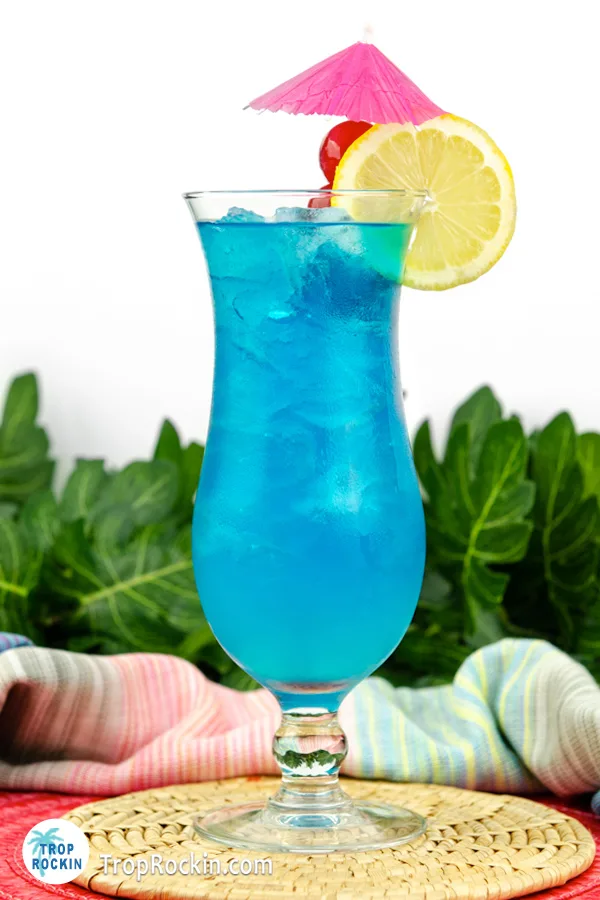 Blue Lagoon Cocktail with Pink Drink Umbrella and Lemon and Cherry Garnish on table with tropical leaves in the background.