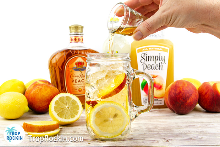 Pouring a shot of Crown Peach whisky into a mason jar filled with ice and fresh peach slices and fresh lemon slices.