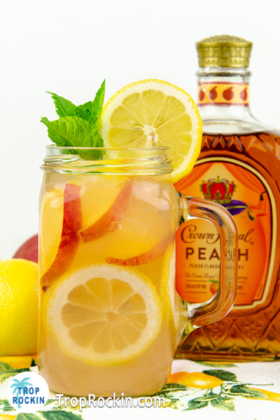 Crown Peach Lemonade in a mason jar with fresh peach slices and lemon wheels and a bottle of Crown Peach in the background.