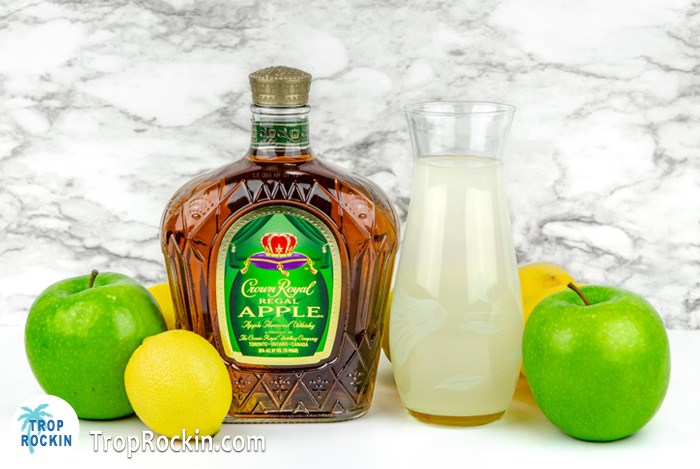 Crown Royal Apple Bottle and caraft of lemonade with fresh apples and lemons on counter top.