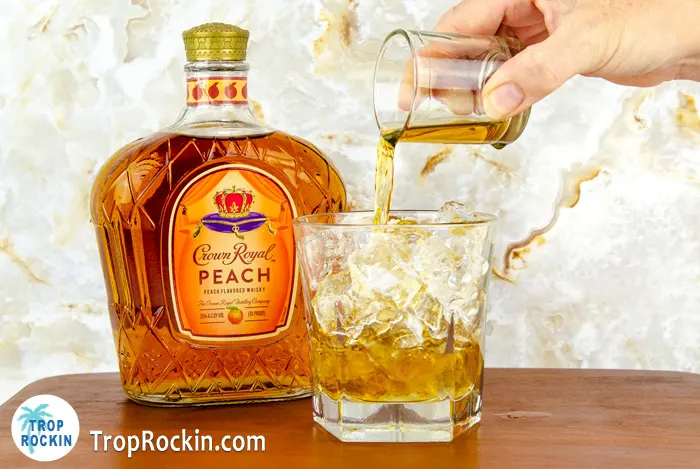 Pouring a shot of Crown Royal Peach Whiskey into a whisky glass filled with ice with Crown Peach bottle in background.