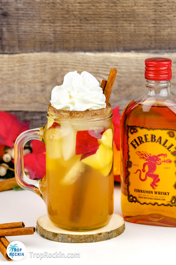 Chilled Fireball Apple Cider drink with ice, whipped cream topping and bottle of Fireball Whiskey in background.