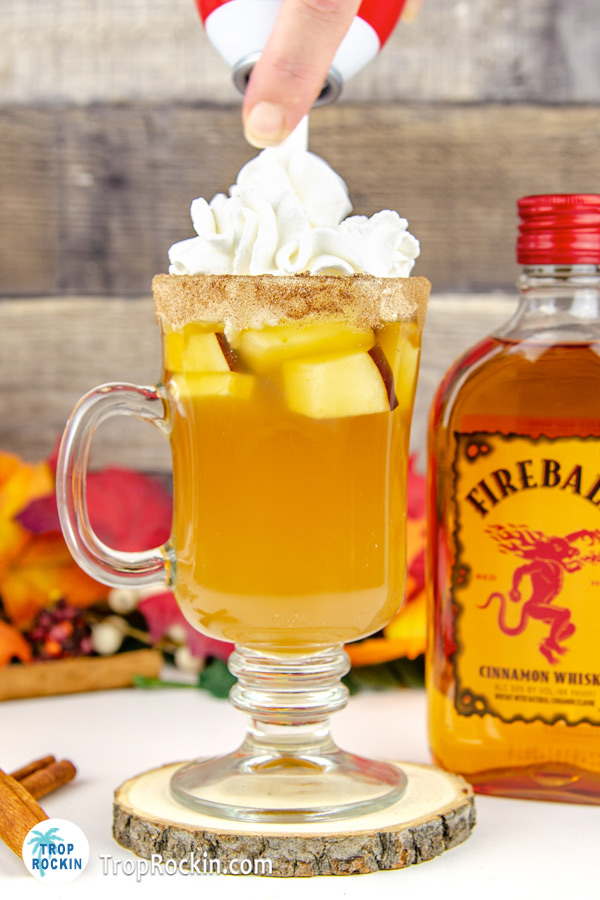 Fireball Apple Cider drink adding whipped cream topping and bottle of Fireball Whiskey in background.