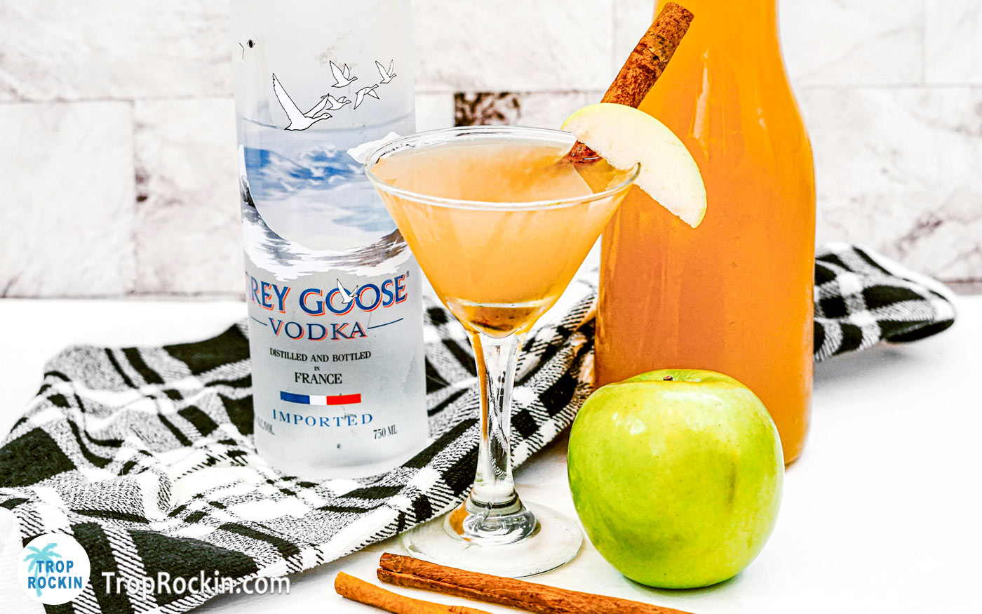 Apple Cider Martini with a fresh slice of apple and a cinnamon stick for garnish. Grey Goose Vodka bottle and caraft of apple cider in background.