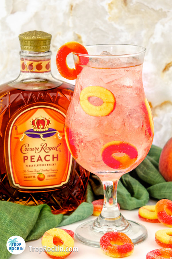 Crown Royal Peach Ring Drink in a hurricane glass with peach rings for garnish inside the glass with a bottle of Peach Crown in the background.