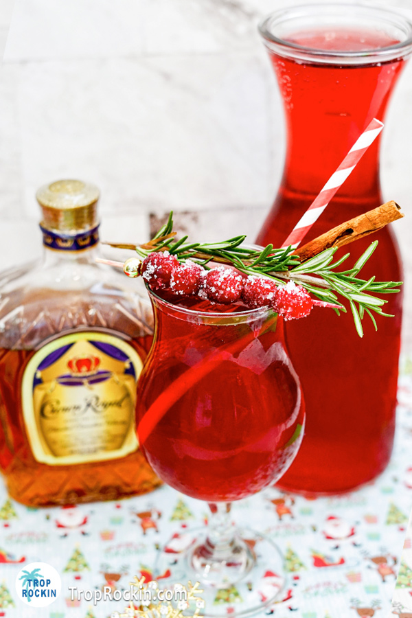 Cranberry Rosemary Cocktail garnished with fresh cranberries and sprig of rosemary with bottle of Crown Royal Whisky and a caraf of cranberry juice in background.