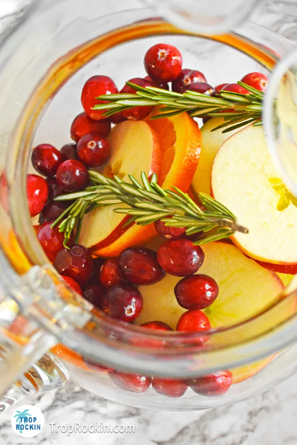 Large pitcher with fresh apple slices, orange slices, fresh cranberries and 2 sprigs of rosemary.