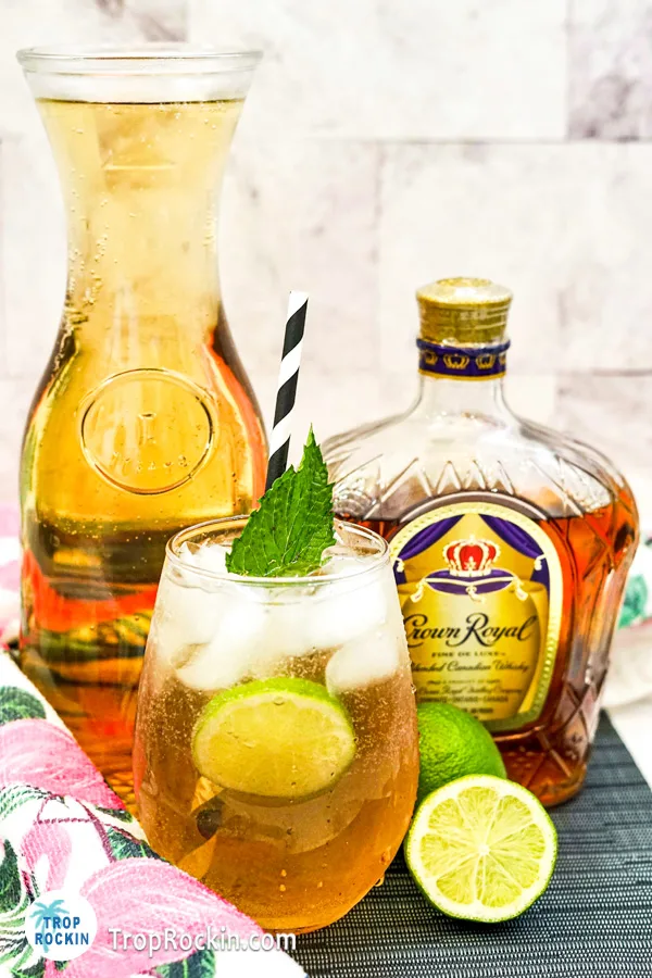Crown Royal and Ginger Ale drink with a slice of lime and sprig of mint for garnish with a caraft of ginger ale and bottle of crown royal in the background.