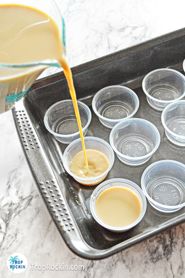 Pouring jello shot mixture into mini jello shot cups which are lined up in a baking pan.