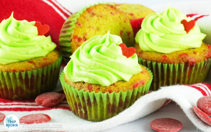 Grinch cupcakes displayed on red and white kitchen towel with red candy melts sprinkled on the counter top.