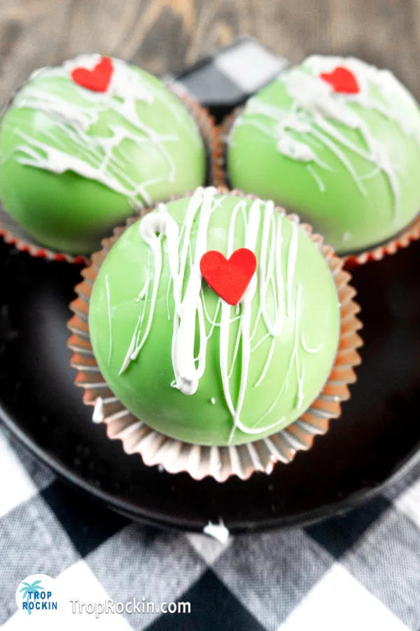 Three Grinch hot chocolate bombs in a blacj bowl with white candy drizzle and a red candy heart on top.