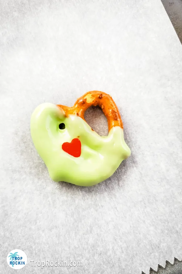 A single Grinch Pretzel dipped in green candy melts with red candy heart on top of the pretzel on a piece of parchment paper.