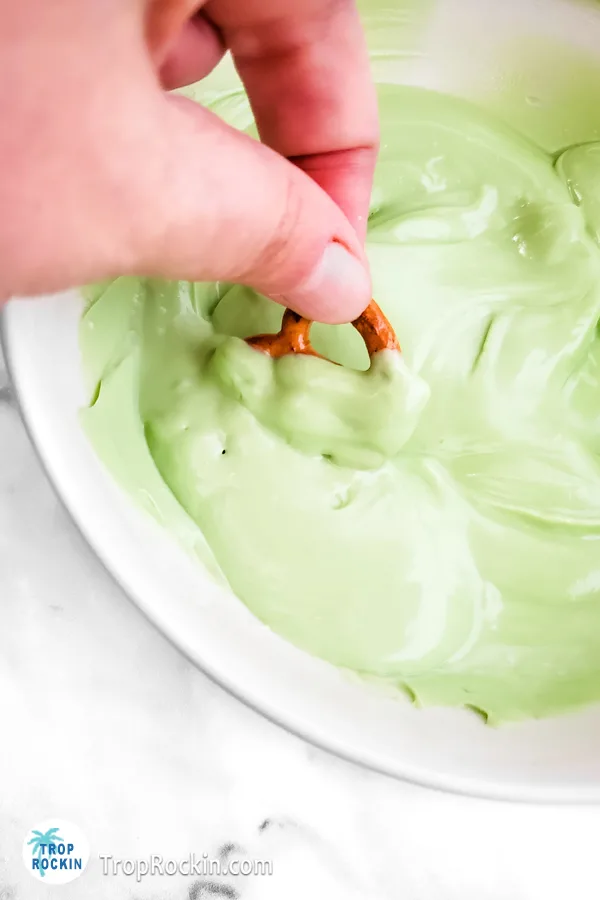 Holding one pretzel and dipping it into a bowl of melted green candy melts.