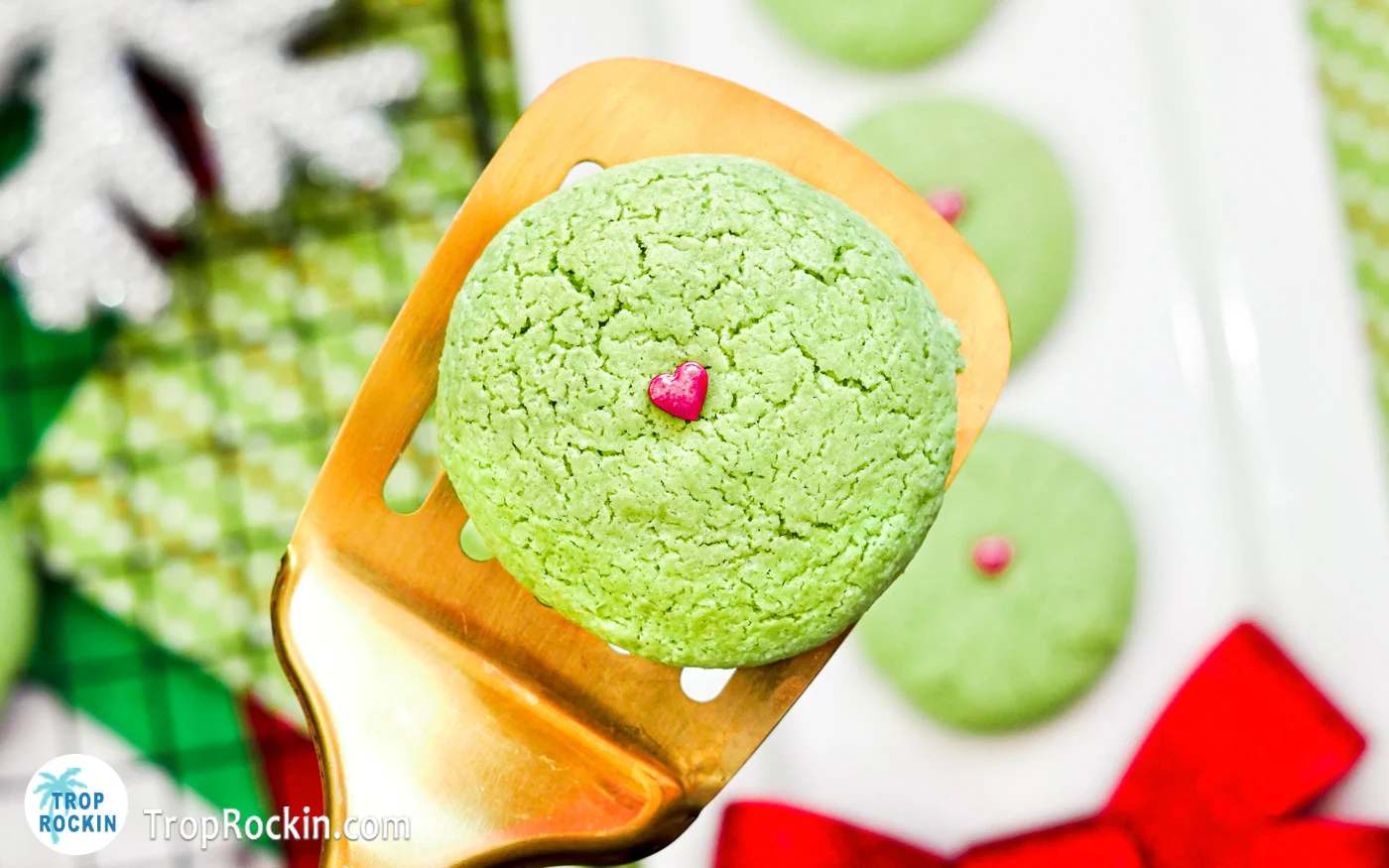 Grinch Sugar Cookies with a red candy sprinkle on top displayed on a spatula.