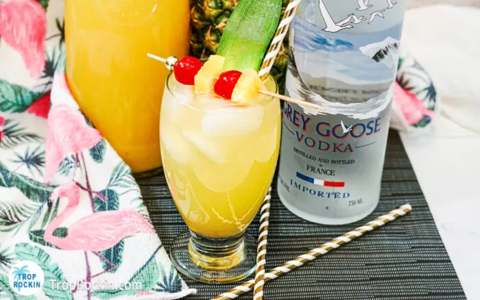 Pineapple Screwdriver with pineapple chunks and Maraschino cherries for garnish on counter top with partial bottle of Grey Goose vodka and a caraft of juice in the background.