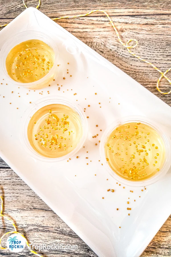 Champagne jello shots with gold sprinkles on a white rectangular serving tray with extra gold sprinkles for decoration.