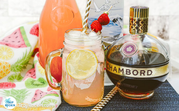 Vodka Raspberry lemonade with lemon wheel and fresh raspberries with caraft of raspberry lemonade, vodka bottle and chambord bottle in background.