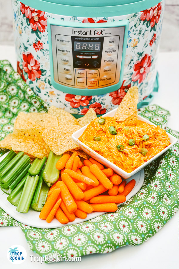 Pressure Cooker Buffalo Chicken Dip in a bowl with a platter of tortilla chips, celery sticks and carrot sticks with the Instant Pot in the background, 