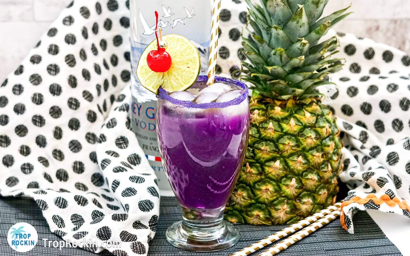 Purple Rain drink with lime and cherry garnish with a fresh pineapple and bottle of vodka in the background.