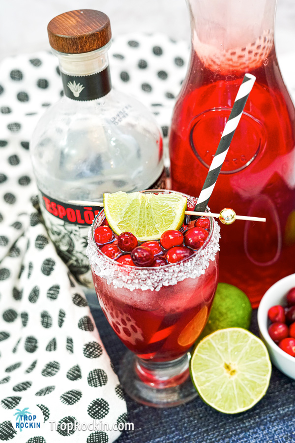 Tequila Cranberry drink topped with fresh cranberies, with a caraft of cranberry juice, bowl of cranberries, sliced limes and bottle of white tequila in the background.