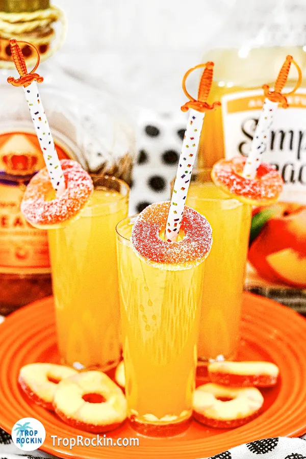 Three Crown Royal Peach Shots with peach rings for garnish on a serving plate with a bottle of crown royal peach and a bottle of peach juice in the background.