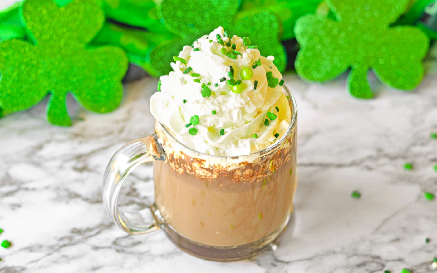 Irish Hot Chocolate in a clear mug topped with whipped cream and St. Patrick's Day sprinkles.