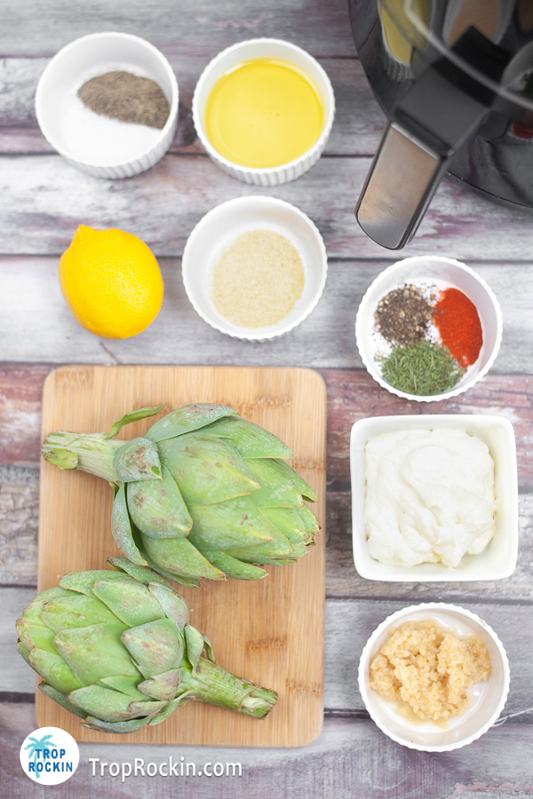 Air fryer artichoke ingredients and garlic aoili suace ingredients displayed in small bowls on counter top with an air fyrer in the background.