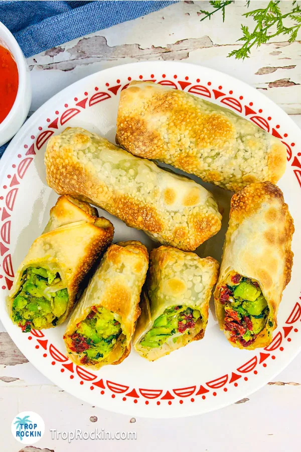 Four avocado egg rolls on a serving plate with 2 cut in half showing the inside avocado filling.