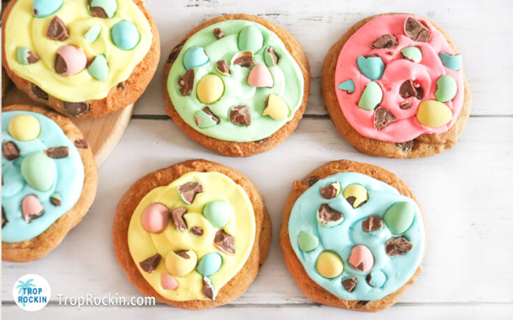 Easter Chocolate Chip Cookies with 4 colors of icing and chopped cabury mini eggs on top displayed on a white wood background.