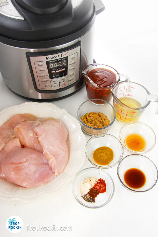 Instant pot shredded BBQ chicken ingredients displayed on counter top in small bowls with raw chicken on a plate. Instant Pot in the background.