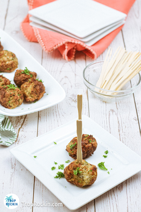 Two air fried chicken meatballs on a small serving plate with forked toothpicks stuck in each one. Background includes a stack of small plates, a ramiken with forked toothpicks and a portion of a white platter filled with chicken meatballs.