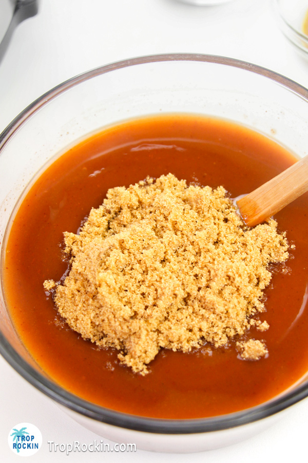 BBQ sauce in a glass bowl with a half cup of brown sugar dumped on top with a wooden spoon in the bowl.