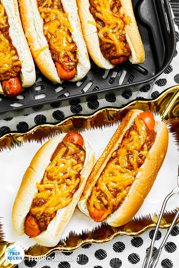 Two Air fryer hot dogs on serving tray with hot dogs in the air fryer basket in the background.