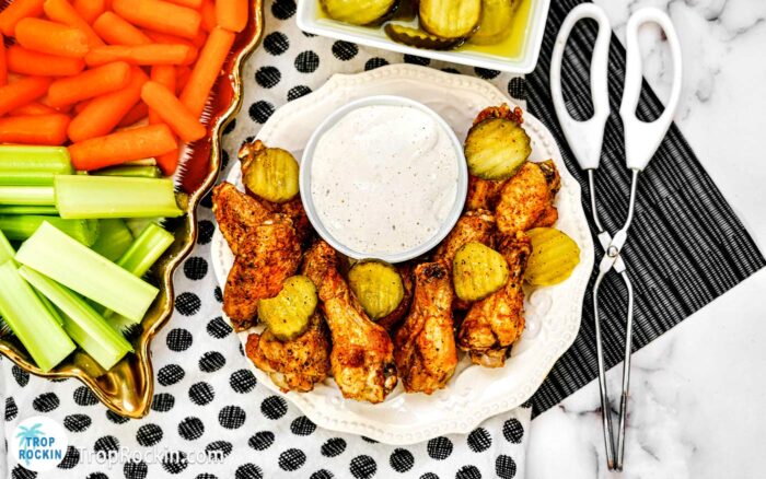Air Fryer Nashville Hot Wings on plate with pickle slices and a bowl of ranch dressing. Serving platters in the background with celery sticks and carrot sticks and a bowl of pickle slices.