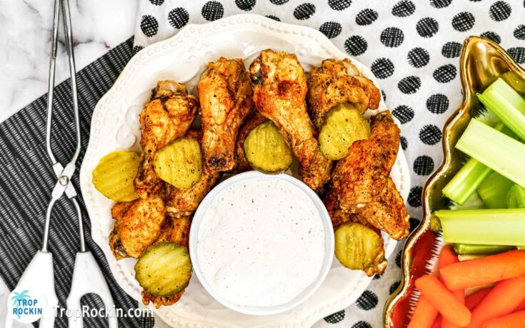 Air Fryer Nashville Hot Wings on plate with pickle slices and a bowl of ranch dressing. Serving platters in the background with celery sticks and carrot sticks and a bowl of pickle slices.