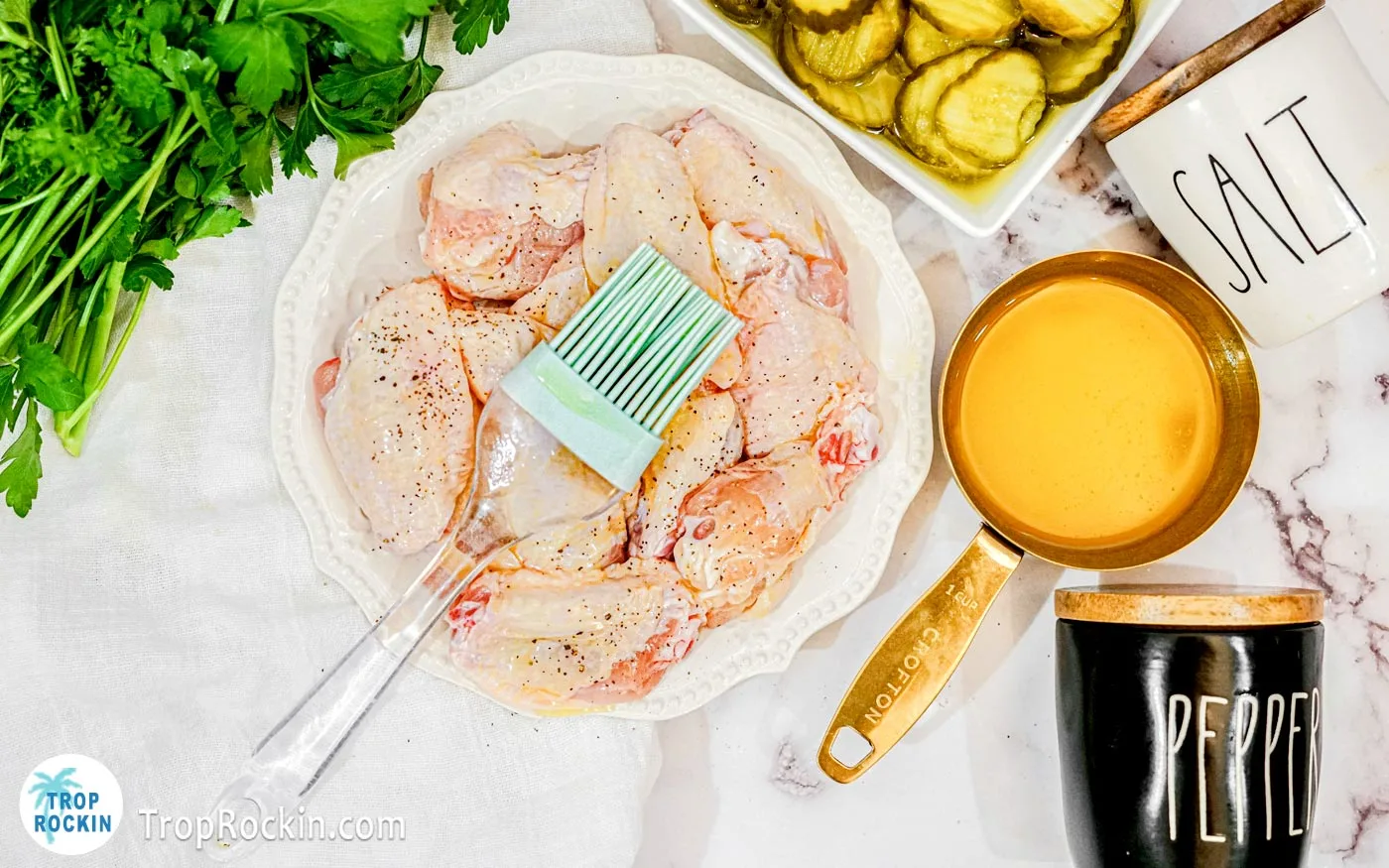 Bowl of raw chicken wings with a pastry brush on to. Small pot of melted butter and bowl of pickle slices next to the chicken wings.