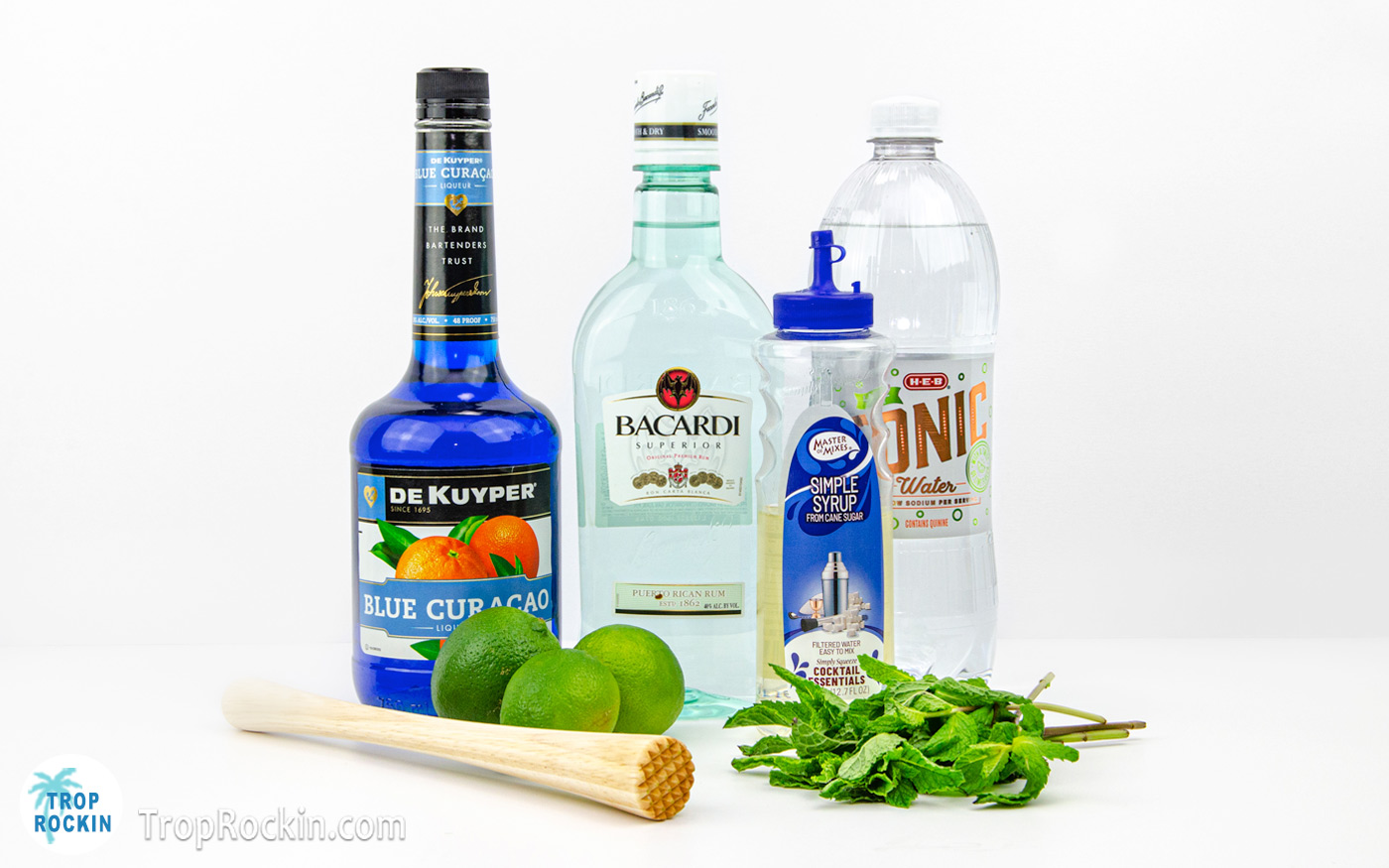 Blue mojito recipe ingredients displayed on a white counter top including a wooden muddler.