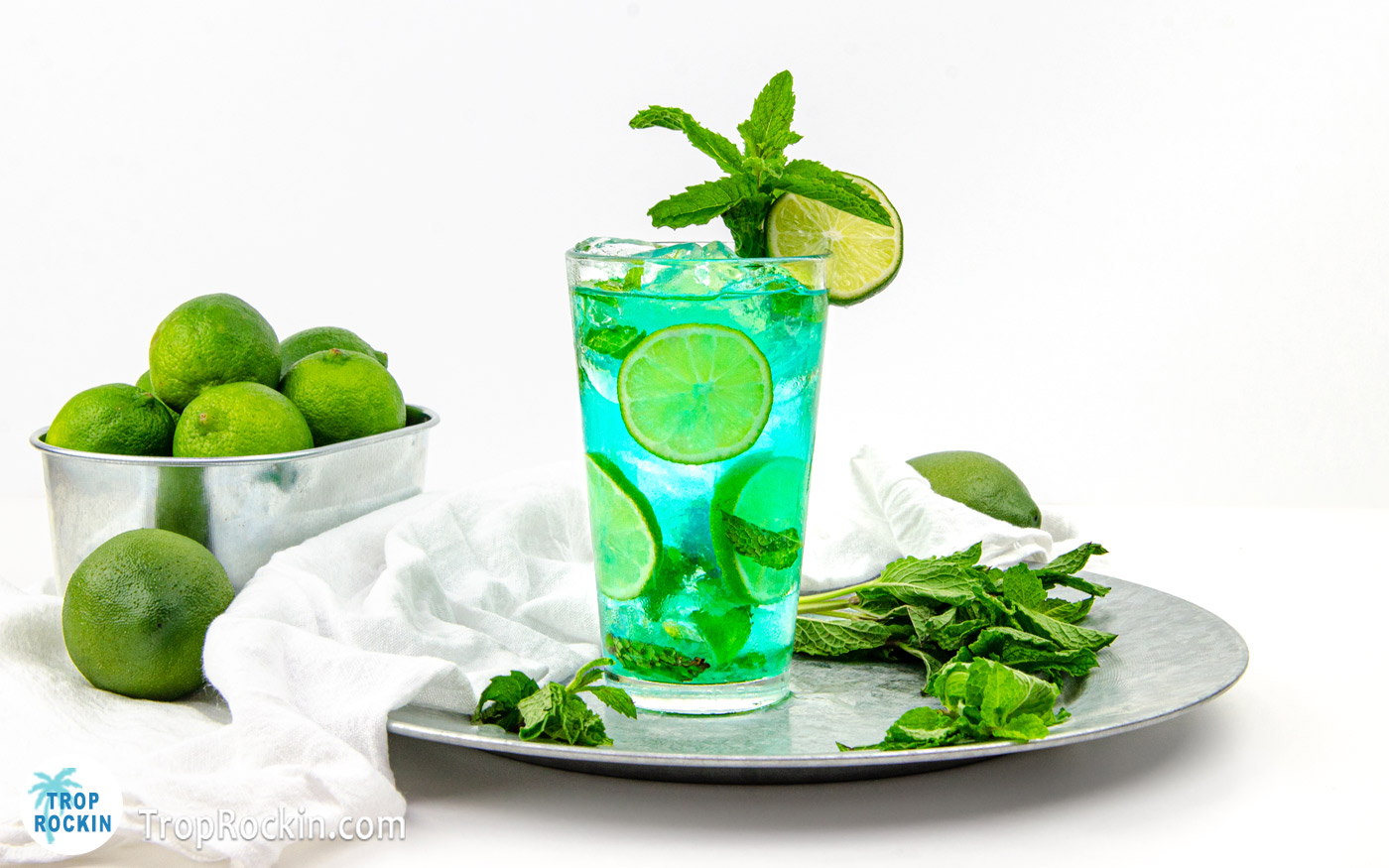 Blue mojito drink with fresh mint leaves and fresh lime wheels in the glass. Garnished with a mint sprig and slice of lime. The drink is sitting on a silver platter with fresh mint scattered around. A metal tub of limes is in the background.