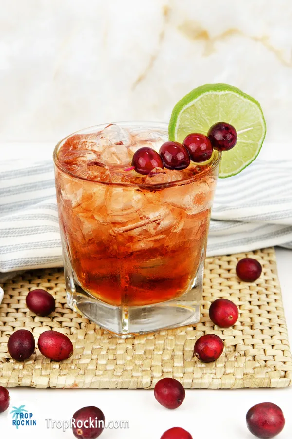 Crown and cranberry drink with four cranberries on a cocktail skewer and a lime wheel for garnish. Sitting on a rattan trivet on a white counter top with fresh cranberries sprinkled around the drink.