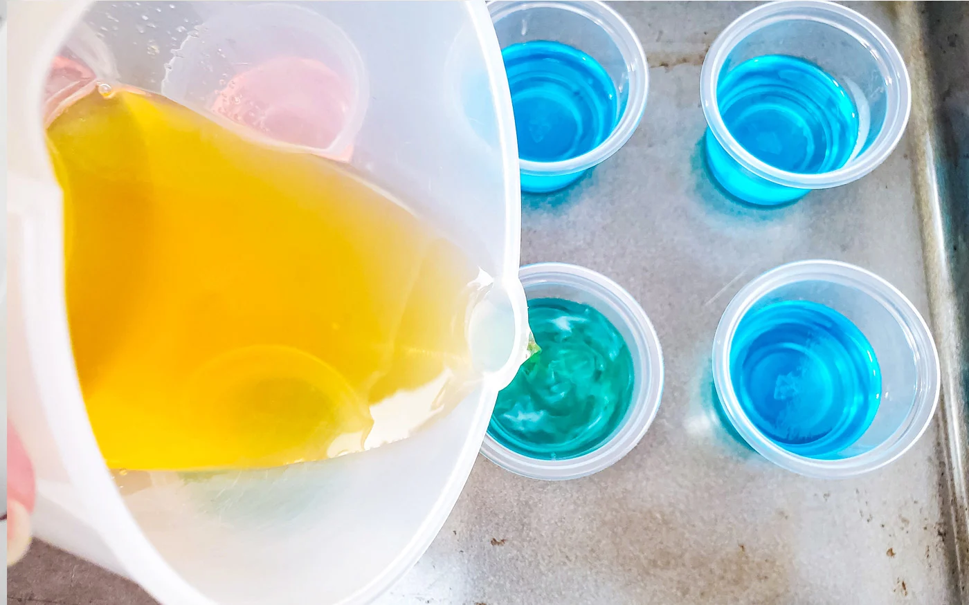 Pouring the lemon starburst jello mixture over the chilled blue raspberry jello in a plastic shot glass sitting on a cookie sheet.