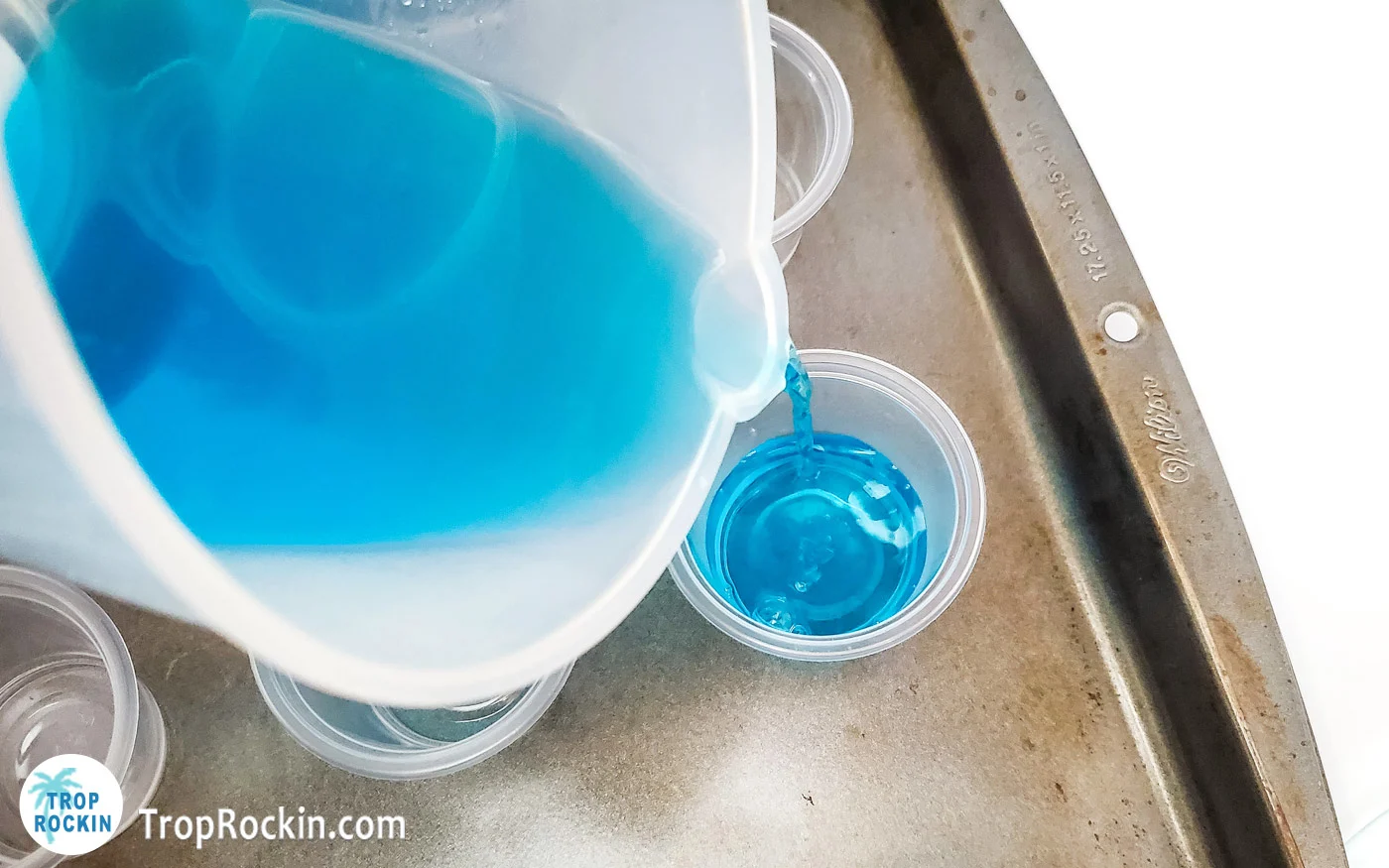 Pouring the blue starburst jello mixture into a mini plastic shot glass sitting on a baking sheet.